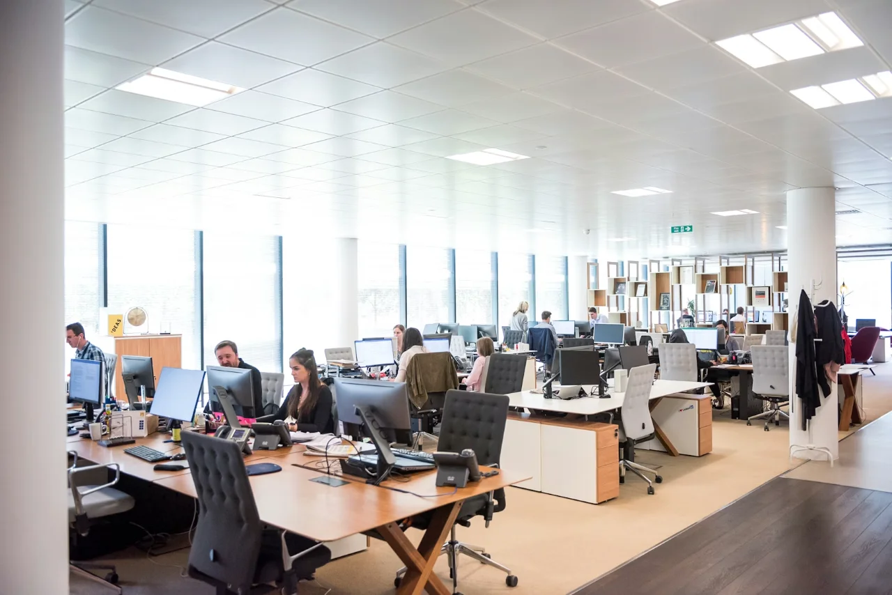 Un espace de bureau ouvert avec de nombreuses personnes travaillant à des bureaux. L'espace est lumineux avec de grandes fenêtres et un mobilier moderne.