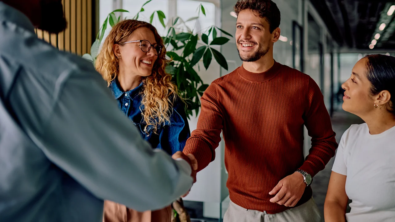 Vier lachende collega's, waarvan twee elkaar de hand schudden in een modern kantoor. Een man in een bruine trui en een vrouw met krullend haar en bril lachen terwijl een man in een grijs overhemd en een vrouw toekijken.